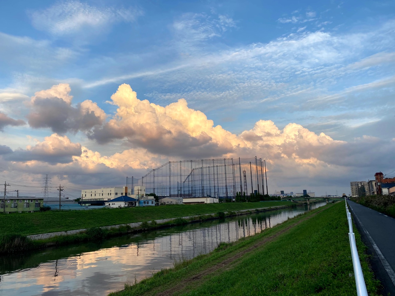 夕景の川口。入道雲が夕日に染まり、川面に映る。土手沿いにサイクリングロードが続く